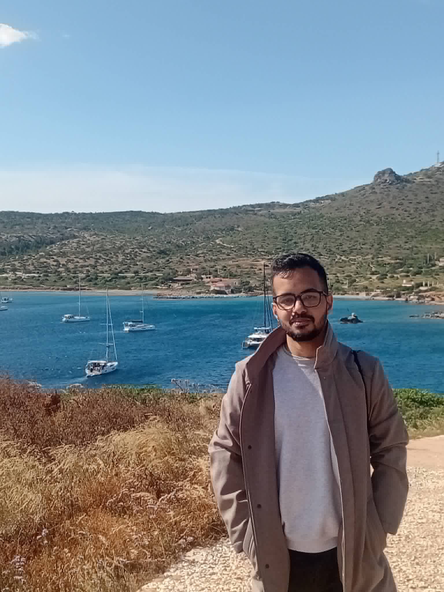 Sudarshan by the sea, with sailboats and hills in the background